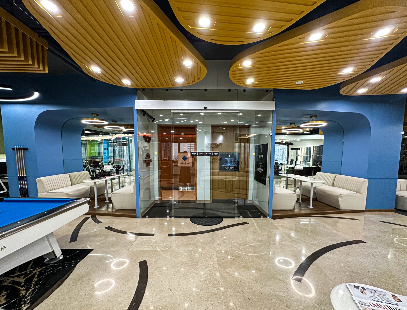 Modern office lounge and recreation area featuring blue accent walls, glass entrance doors, white seating, a billiards table, polished marble flooring, decorative wooden ceiling panels, and contemporary ring lighting.